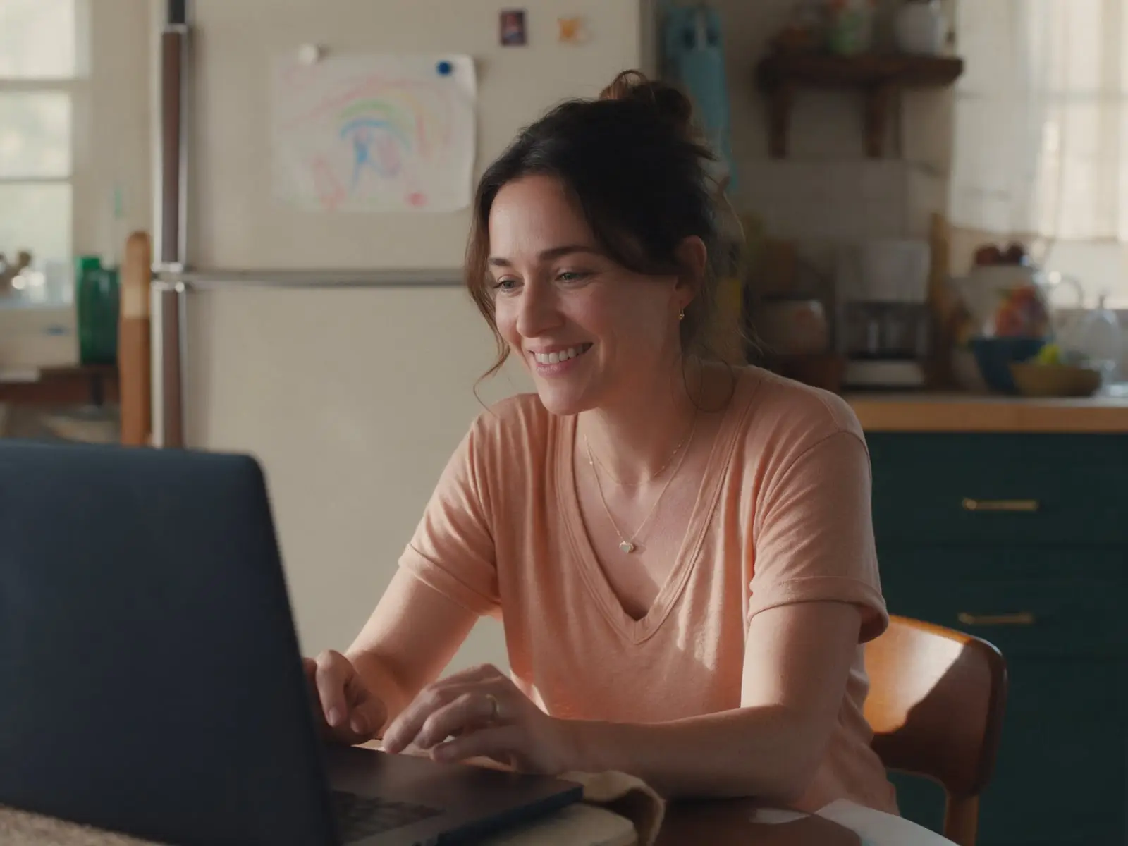A woman with her hair pinned up smiling during a video call at her kitchen table, a child's crayon drawing on the fridge behind her