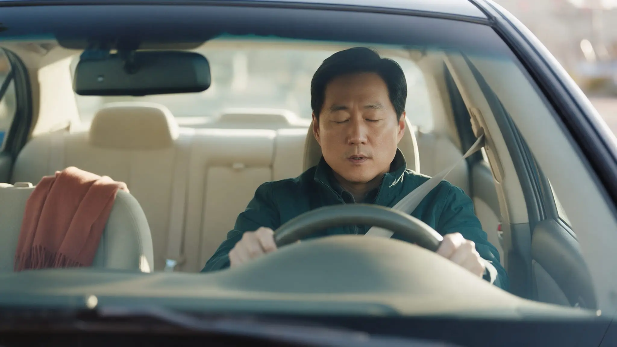 A man in a dark green jacket sitting in a parked car with his seatbelt still on and both hands on the wheel, eyes closed as he takes a breath, a rust-colored scarf on the passenger seat