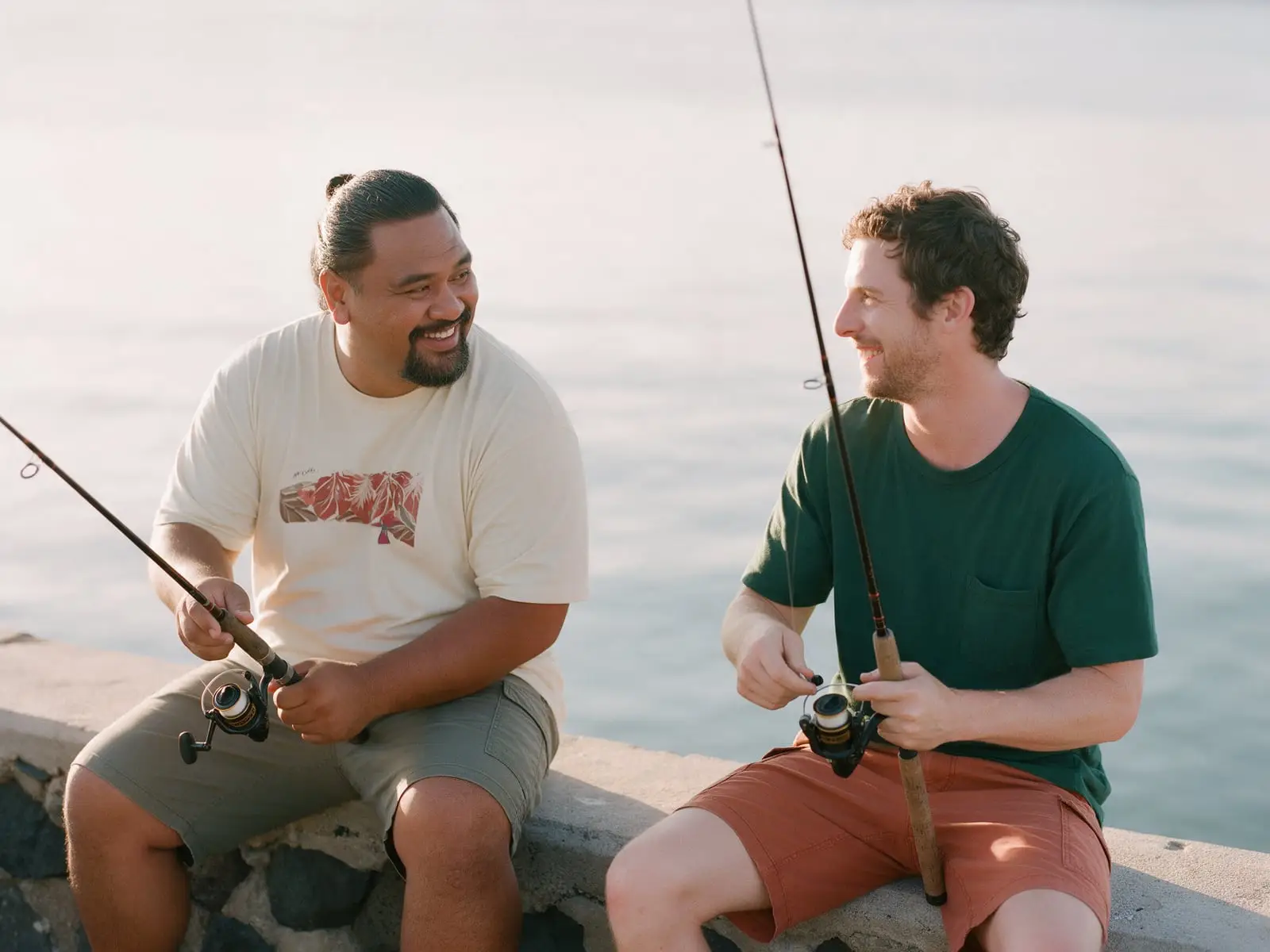 Two friends laughing as they sit fishing on a low seawall on a calm, hazy morning