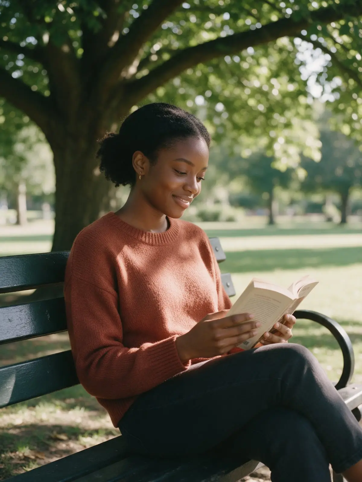 A young woman in a rust-colored sweater reading a paperback on a park bench under a broad shade tree, smiling slightly