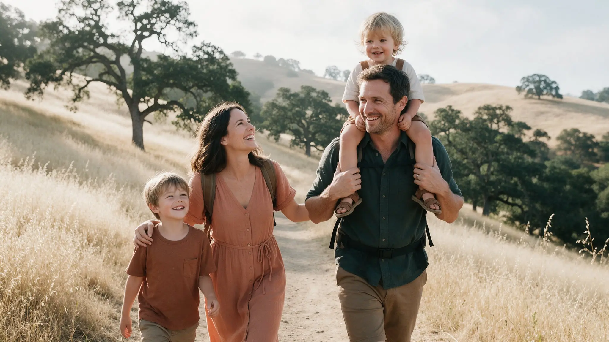 A family of four smiling as they hike a trail through golden grass and oak trees, a small child riding on the father's shoulders
