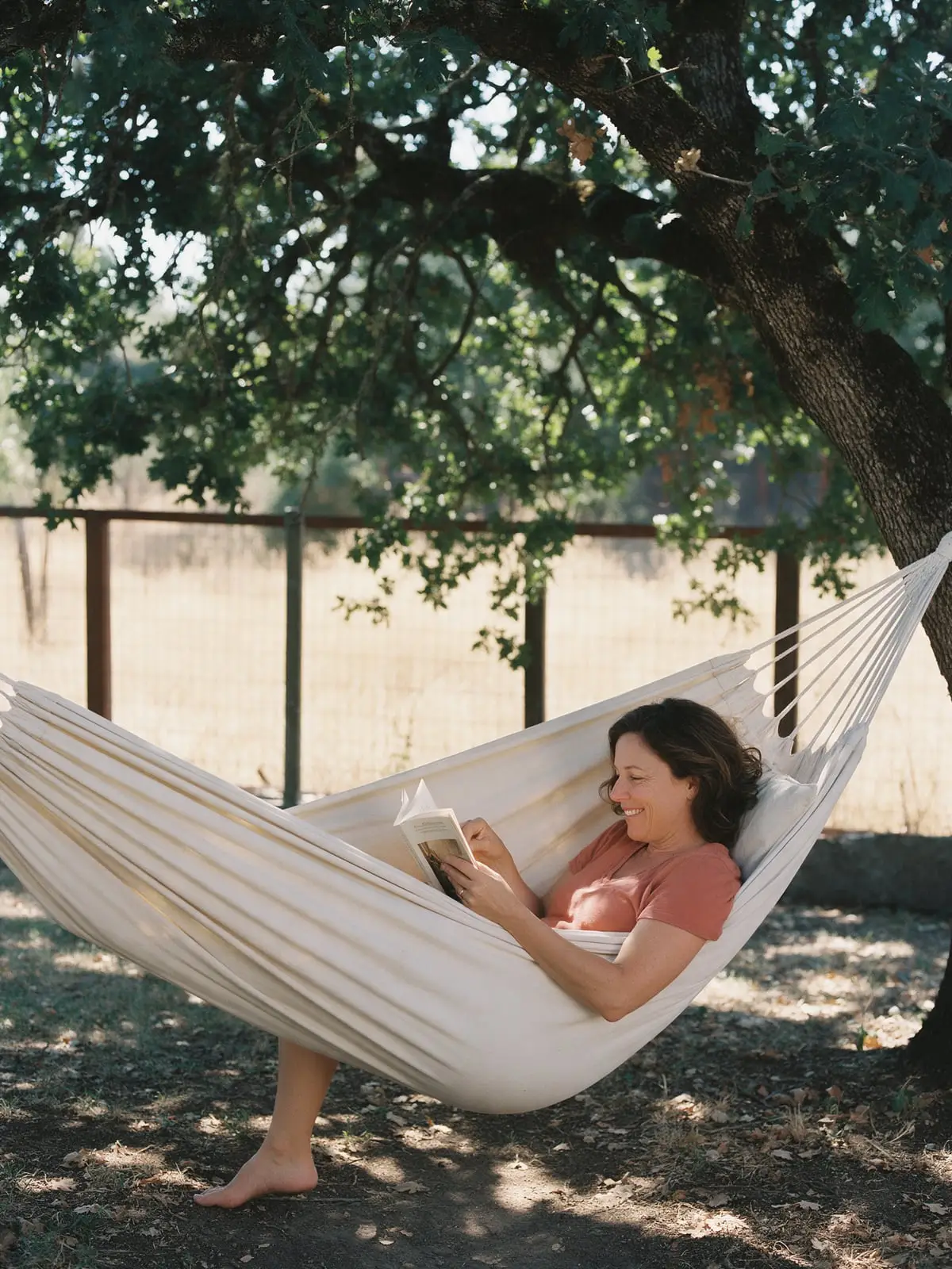 A woman smiling at her book as she lies in a canvas hammock under a large oak tree, dry golden grass beyond the fence