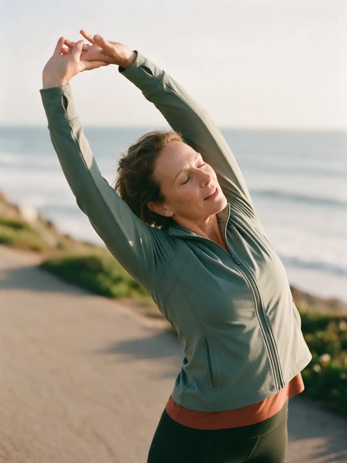 A woman in a sage green jacket stretching her arms overhead on a coastal walking path, the ocean soft behind her
