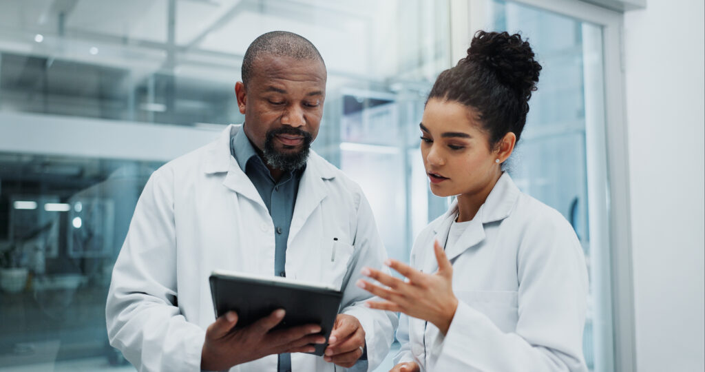 Two scientists in lab coats review a tablet in a glass-walled laboratory.