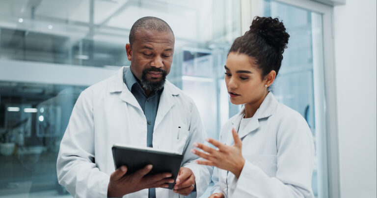 Two scientists in lab coats review a tablet in a glass-walled laboratory.
