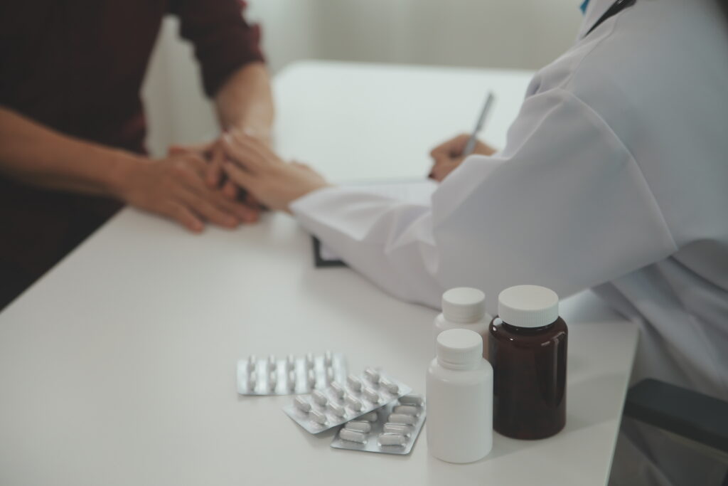 Medical professional in a white coat writing at a table with pill bottles and blister packs nearby, blurred background of a patient.