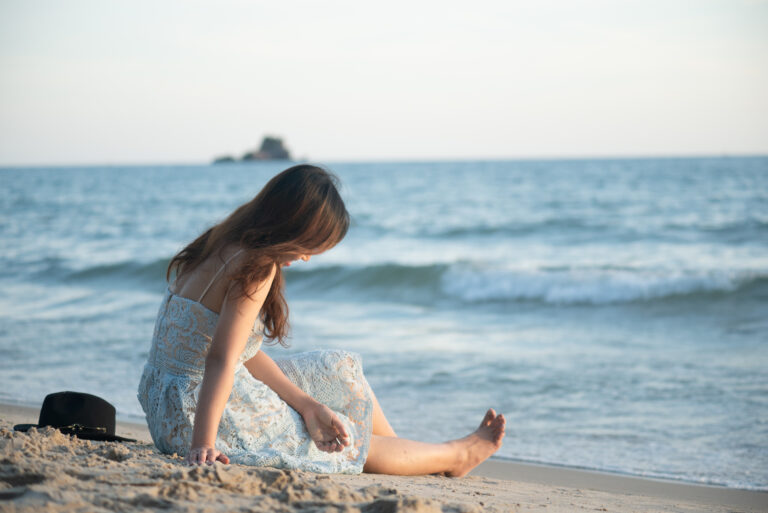 A girl in a white lace dress sits on the sandy beach, toes in the sand, with the ocean and distant horizon behind her.